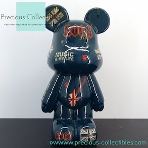 Peut inclure: Une figurine d'ours en céramique bleu foncé avec une finition brillante. L'ours a de grandes oreilles et est décoré de divers motifs colorés, dont le drapeau de l'Union Jack, des notes de musique et les mots "Rock Star" et "Music is my life".