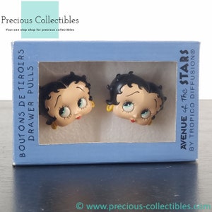 May include: A pair of drawer pulls featuring the cartoon character Betty Boop. The pulls are made of plastic and are painted in a realistic style. The packaging is blue and features the text "Boutons de Tiroirs Drawer Pulls Avenue of The Stars by Tropico Diffusion".