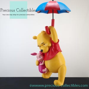 Könnte beinhalten: Figur Winnie Puuh und Ferkel. Puuh hält Ferkel und beide hängen an einem roten und blauen Regenschirm. Die Figur ist aus gelbem, rotem und rosa Kunststoff.