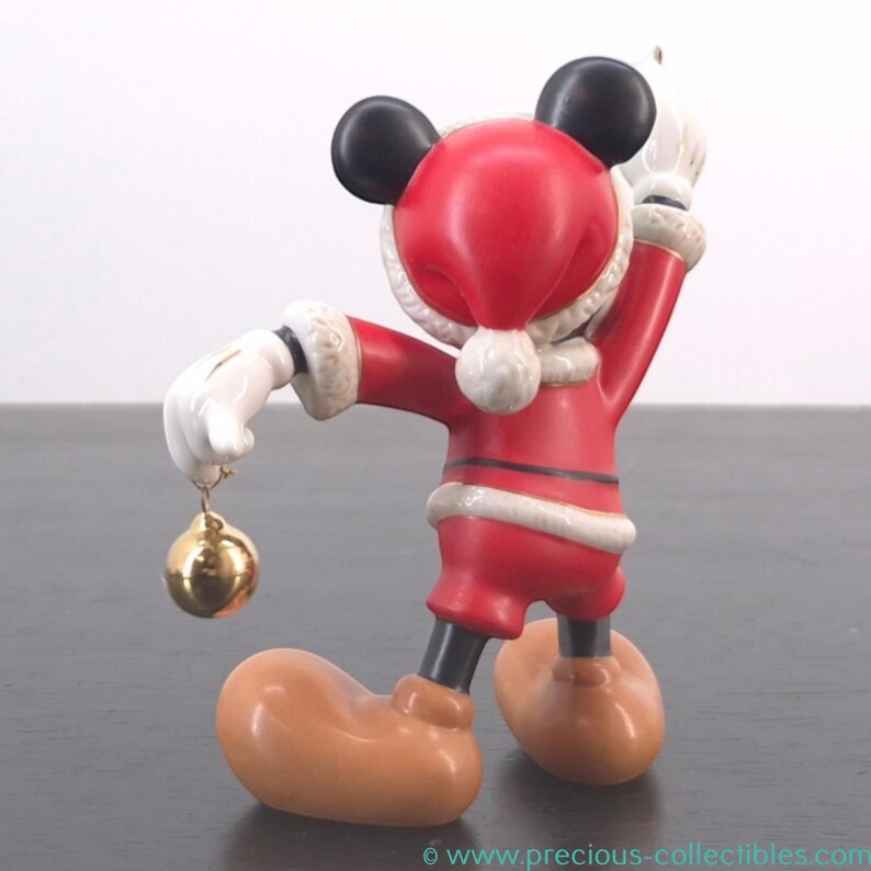May include: A figurine of a cartoon mouse dressed in a Santa Claus outfit. The red suit has white trim and a matching hat. The mouse is holding a gold ornament in one hand and has black ears and large tan shoes.