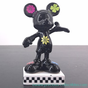 May include: A black Mickey Mouse figurine with colorful floral and clover accents. The ears feature a pink flower and a green four-leaf clover. A yellow flower adorns the chest, and the feet have red and blue details. The figurine stands on a white base with a black and white checkered pattern.