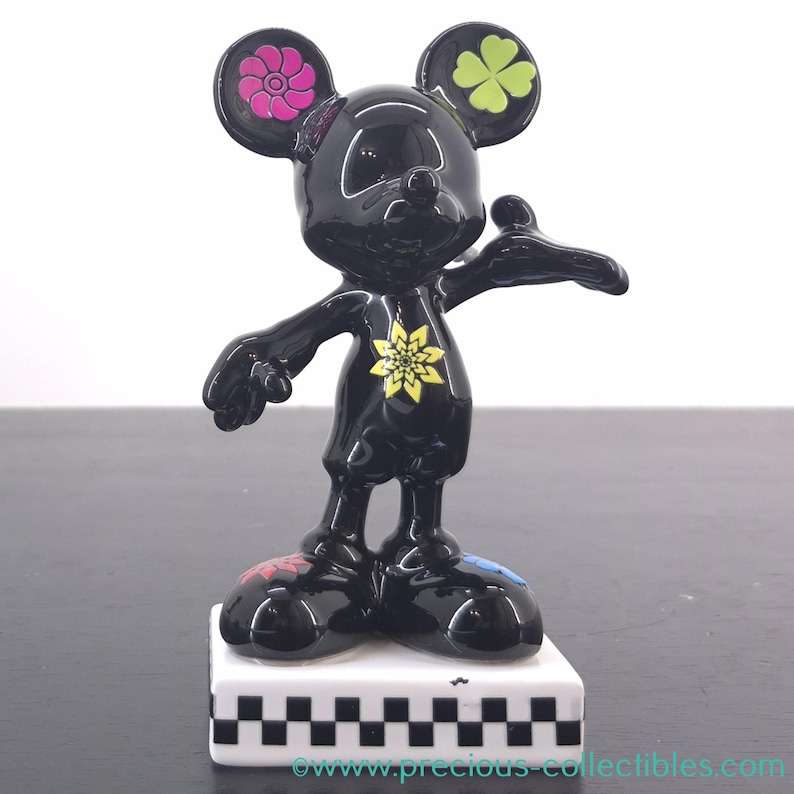 May include: A black Mickey Mouse figurine with colorful floral and clover accents. The ears feature a pink flower and a green four-leaf clover. A yellow flower adorns the chest, and the feet have red and blue details. The figurine stands on a white base with a black and white checkered pattern.