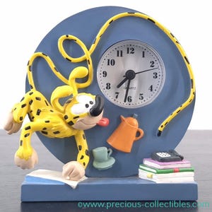 Puede incluir: Un reloj de figura de leopardo de dibujos animados azul y amarillo con una esfera blanca y agujas negras. El reloj está rodeado de una cuerda amarilla y tiene una cafetera, una taza y un teléfono sobre una pila de libros.