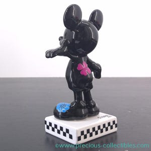 May include: A black Mickey Mouse figurine with a glossy finish. The figure has a pink flower on its chest and a blue flower on one foot. It stands on a white base with a black and white checkered pattern.