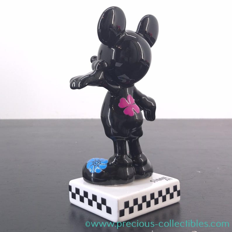 May include: A black Mickey Mouse figurine with a glossy finish. The figure has a pink flower on its chest and a blue flower on one foot. It stands on a white base with a black and white checkered pattern.