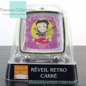 Puede incluir: Un reloj despertador cuadrado verde con una imagen de dibujos animados de Betty Boop con un atuendo rosa y amarillo. La esfera del reloj tiene números y manecillas negros. El reloj está empaquetado en una caja de plástico transparente con una base negra que dice "Avenue of the Stars" y "Réveil Retro Carré".