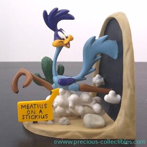 May include: A Road Runner ornament with a purple head crest, blue body, and yellow beak. The ornament is set in a desert scene with a sign that reads "MEATIUS ON A STICKIUS".