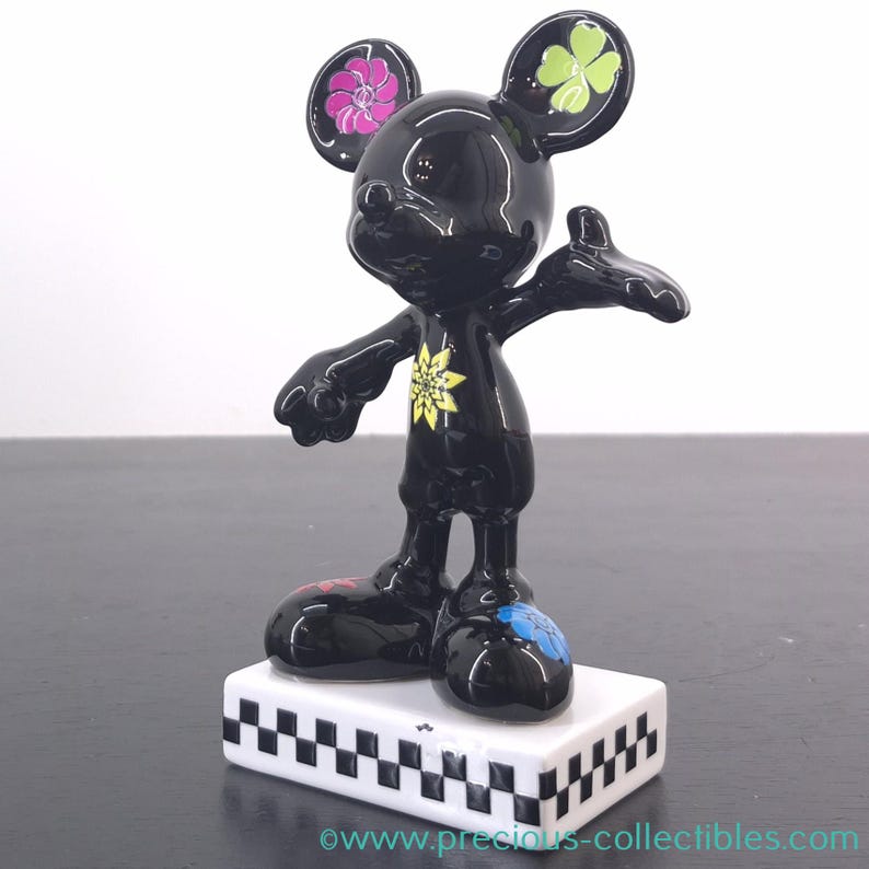 May include: A black Mickey Mouse figurine with floral and clover accents on the ears. The figurine has a yellow flower design on the chest and stands on a white base with a black and white checkered pattern.