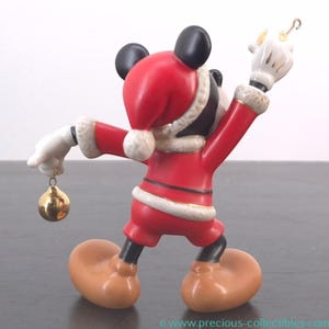 May include: A figurine of Mickey Mouse dressed in a Santa Claus outfit. The red suit has white trim and a matching hat. Mickey is holding a gold ornament in one hand and a gold hook in the other. The figurine has black ears and a tan face and feet.
