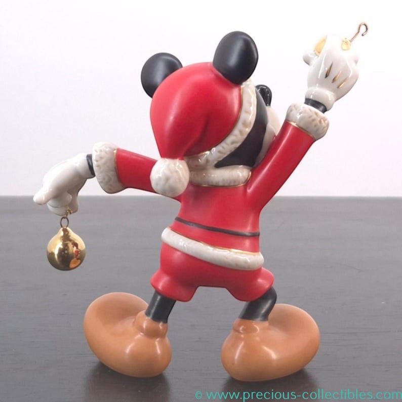 May include: A figurine of Mickey Mouse dressed in a Santa Claus outfit. The red suit has white trim and a matching hat. Mickey is holding a gold ornament in one hand and a gold hook in the other. The figurine has black ears and a tan face and feet.