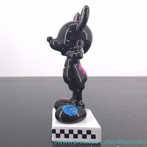 May include: Black figurine of a mouse-like character with large ears, standing on a white base. The figurine has pink and yellow floral accents and blue flowers on the feet. The base has a black and white checkered pattern.