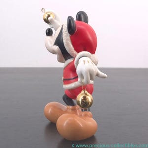 May include: A Mickey Mouse figurine dressed in a Santa Claus outfit, holding two gold Christmas ornaments. The figurine has a red coat with white trim, black ears, and white gloves. The figure is standing on a dark surface.