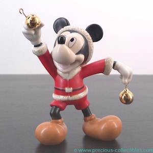May include: A Mickey Mouse figurine dressed in a Santa Claus outfit, holding two gold Christmas ornaments. The figurine is wearing a red suit with white trim, a black head, and tan shoes. The figurine is a collectible item.