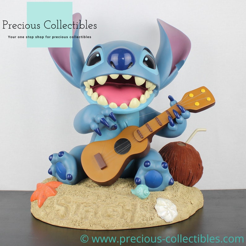 Puede incluir: Figurilla azul de Stitch tocando un ukelele mientras est&aacute; sentado en una playa con un coco y una estrella de mar. La figurilla est&aacute; sonriendo y tiene una expresi&oacute;n feliz.