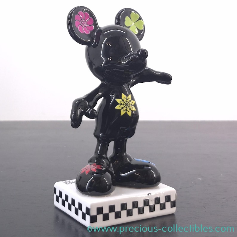 May include: A black Mickey Mouse figurine with floral accents on the ears, chest, and feet. The figurine stands on a white base with a black and white checkered pattern. The figurine is a collectible item.