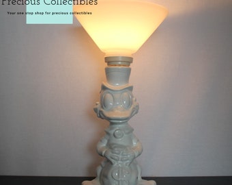 Può includere: Lampada in ceramica bianca con una statuetta di anatra che tiene un sacco di soldi. Il paralume è bianco e ha una forma larga e arrotondata. L'anatra indossa un cappello a cilindro e ha un becco e occhi grandi. Il sacco di soldi ha un segno di dollaro. La lampada è su una superficie scura.