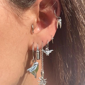 Puede incluir: Una colección de pendientes plateados, que incluye aros, un diente, un pájaro, un candado, un murciélago, una daga y una araña. Los pendientes se lucen en una oreja, mostrando una variedad de diseños únicos y detallados.