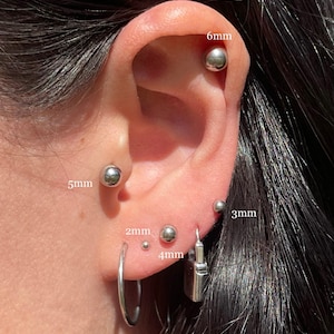 Puede incluir: Primer plano de la oreja de una persona con varios piercings de bola plateados. Los piercings están etiquetados con sus tamaños en milímetros: 6 mm, 5 mm, 4 mm, 3 mm y 2 mm. También hay un pendiente de aro plateado y un pendiente de candado plateado.