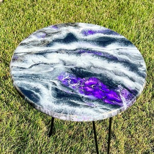 May include: A round, black and white epoxy resin table top with purple accents. The table top is supported by three black metal hairpin legs.