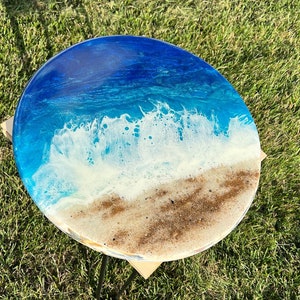 May include: Round table top with a resin ocean scene. The table top features blue water, white foamy waves, and a sandy beach.