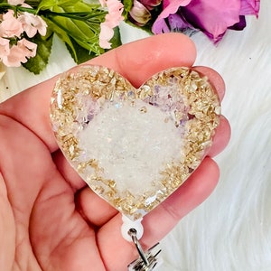 May include: A white heart-shaped resin badge holder with gold glitter and gold leaf accents. The holder is attached to a white retractable badge reel.