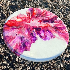 May include: A round side table with a white top featuring a vibrant abstract design in shades of red, pink, and purple. The table has three black metal hairpin legs. The table's diameter is approximately 18 inches.