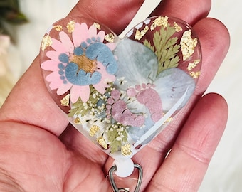 May include: A heart-shaped resin keychain with a floral design and baby footprints. The resin is clear and the flowers are pressed inside. The keychain is attached to a silver retractable badge holder.