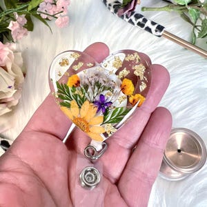 May include: A heart-shaped badge reel with a clear resin design. The resin encases dried yellow and purple flowers, green leaves, and gold flakes. The reel has a silver-colored clip and a clear plastic strap.
