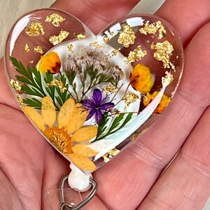 May include: Heart-shaped badge reel with a clear resin design. The badge reel features dried yellow and purple flowers, green leaves, and gold flakes. The badge reel has a white backing and a silver-colored metal clip.