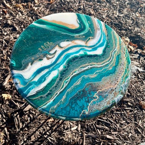 May include: Round decorative art piece with an abstract design. The artwork features flowing patterns of teal, white, gold, and dark green. The piece is circular and appears to be made of resin or a similar material.
