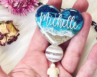 May include: A heart-shaped badge reel with a beach scene design, featuring the name "Michelle" in white script. The reel includes a striped bead, a turtle charm, and a faceted teal bead.