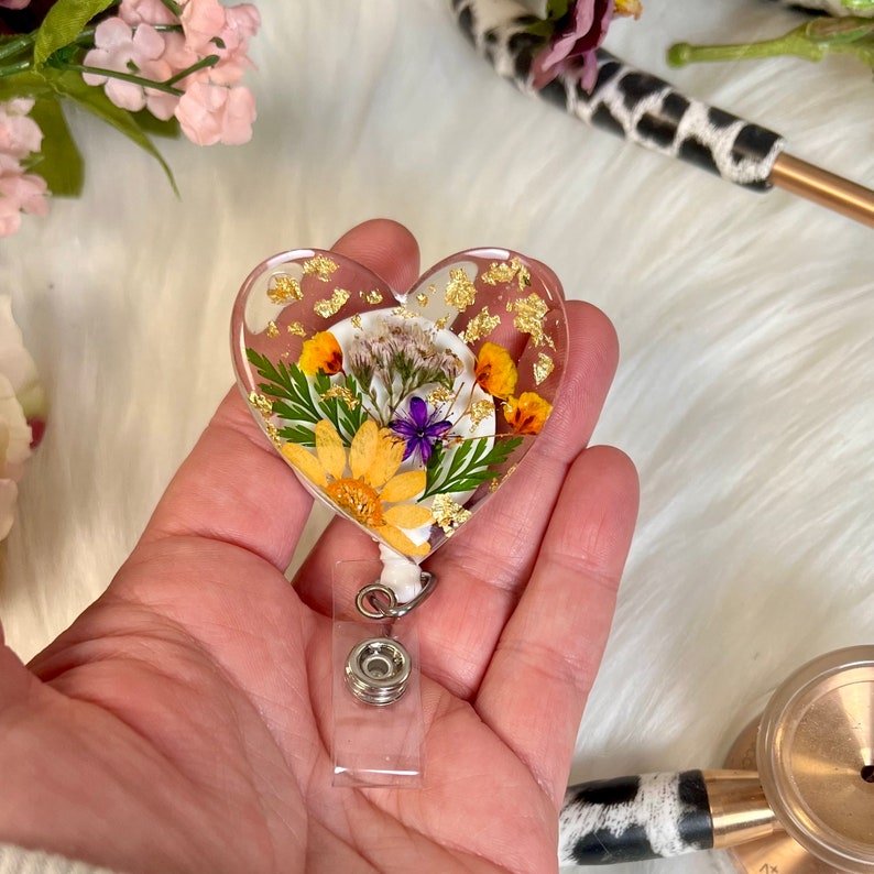 May include: Heart-shaped badge reel with embedded dried flowers and gold flakes. The clear resin design features yellow, purple, and white flowers. The badge reel has a retractable cord and a snap-on clip for attaching to clothing.