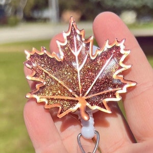 May include: A gold glitter maple leaf with a copper outline is attached to a retractable badge holder. The leaf is a fall-themed design.