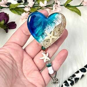 May include: A heart-shaped resin keychain with a blue and white ocean scene, sand, and a seashell. The keychain has a white starfish charm and green and gold beads.
