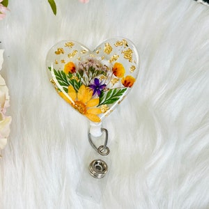 May include: A heart-shaped badge reel with a clear resin design. The resin encases dried yellow and purple flowers, green leaves, and gold flakes. The reel has a retractable cord and a metal clip for attaching to clothing.