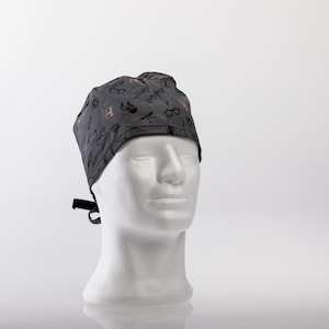 Op de afbeelding: Grijze chirurgische scrub-muts met een zwarte en gouden Harry Potter-print. De print bevat brillen, een bliksemschicht, een toverstaf en de letter H.