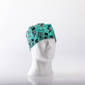 Op de afbeelding: Een turquoise blauwe chirurgische scrub-muts met een kleurrijk patroon van medische instrumenten en harten. De muts zit op een wit mannequin-hoofd.