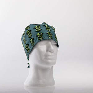 Op de afbeelding: Een blauwe chirurgische scrub-muts met een herhalend patroon van zwarte silhouetten en de tekst "I am the danger" in groen.
