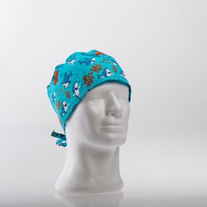 May include: A turquoise blue surgical cap with a pattern of cartoon sharks and coral. The cap has a tie at the back.