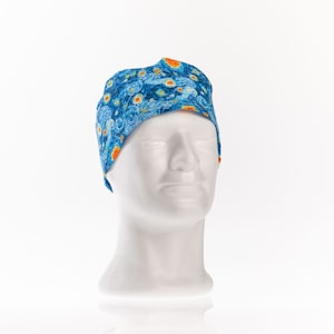 May include: A blue surgical scrub cap featuring a print inspired by Van Gogh's "Starry Night" painting. The cap has a curved design with orange accents and is displayed on a white mannequin head.