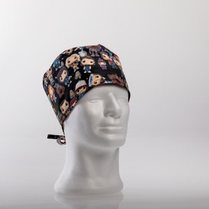 Op de afbeelding: Zwarte scrub-muts met een kleurrijke print van cartoonfiguren. De muts heeft een strik aan de achterkant.