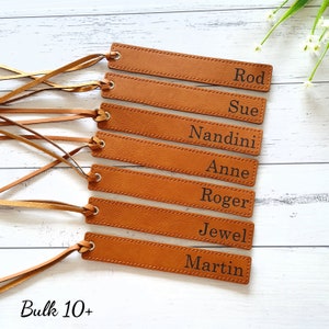 May include: Seven brown leather bookmarks with different names engraved on them. The names are Rod, Sue, Nandini, Anne, Roger, Jewel, and Martin. The bookmarks are arranged in a vertical stack, with the top bookmark at the top of the image and the bottom bookmark at the bottom of the image. The bookmarks are all the same size and shape, and they have a tassel at the top. The bookmarks are on a light wood background. The text "Bulk 10+" is at the bottom of the image.
