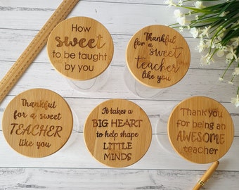 May include: Four round wooden jar lids with engraved messages for teachers. The messages are: "How sweet to be taught by you", "Thankful for a sweet teacher like you", "It takes a BIG HEART to help shape LITTLE MINDS", and "Thank you for being an AWESOME teacher".