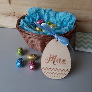 May include: A light blue wicker basket filled with colorful foil-wrapped chocolate eggs. A blue ribbon is tied around the basket and attached to a wooden egg-shaped tag with the name "Max" engraved on it.