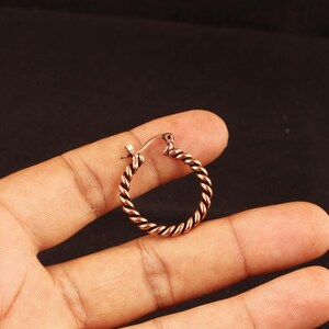 May include: A small, circular hoop earring crafted from copper-toned metal with a distinct twisted rope texture. The earring features a hinged closure mechanism at the top, allowing it to open and close securely for placement through an earlobe.