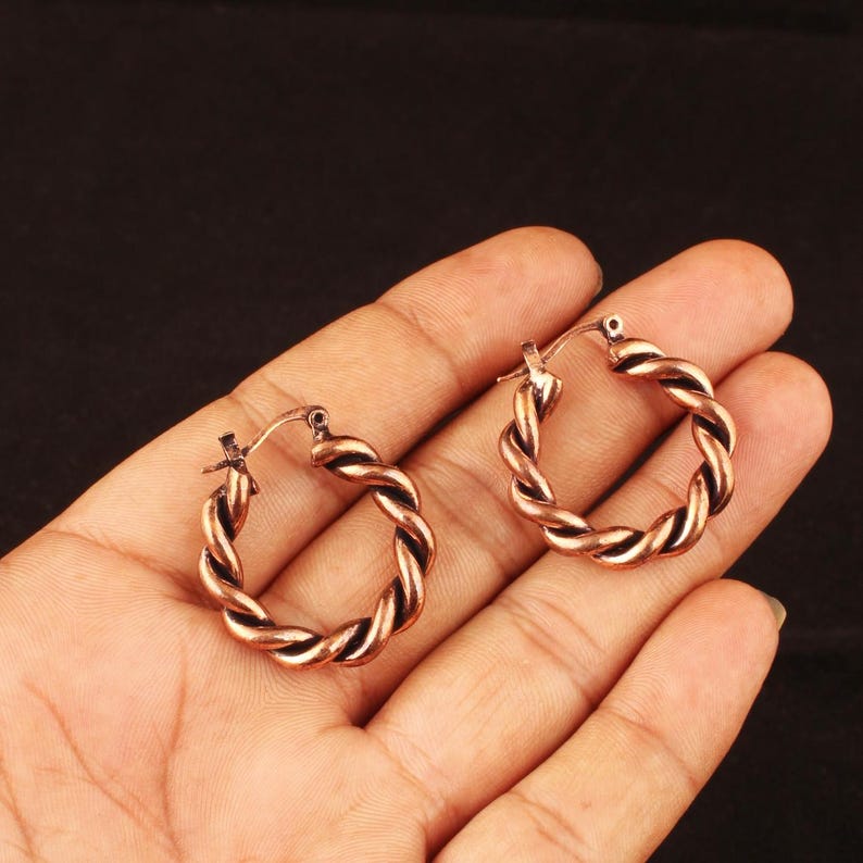 May include: A pair of copper-toned hoop earrings featuring a twisted rope design. Each circular earring has a polished metallic finish with dark recessed areas that highlight the spiral texture of the metal. The hoops include a hinged post closure.
