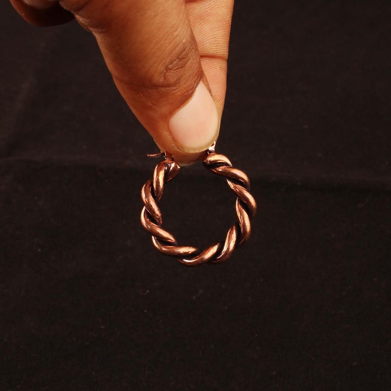 May include: A circular hoop earring crafted from polished copper metal, featuring a distinct twisted rope texture throughout the entire band. The earring has a metallic reddish-brown finish and a simple hinged closure mechanism at the top.