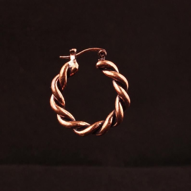 May include: A single hoop earring crafted from a metallic copper-toned material, featuring a distinct twisted rope design. The circular earring includes a hinged post mechanism at the top, allowing it to open and close securely for wear.