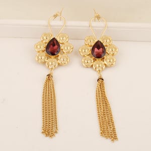 Può includere: Un paio di orecchini color oro con una gemma rosso scuro a forma di goccia al centro, circondata da sei piccoli fiori in metallo. Ogni orecchino presenta una lunga nappa pendente composta da sottili catenelle in metallo color oro.