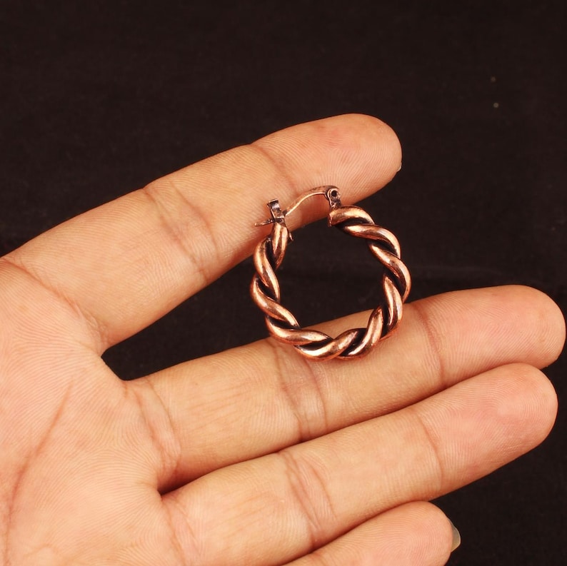 May include: A small, circular hoop earring with a twisted, rope-like texture and a dark, antiqued copper finish. The earring features a hinged post mechanism at the top, designed for secure closure, showcasing a metallic, reddish-brown tone.